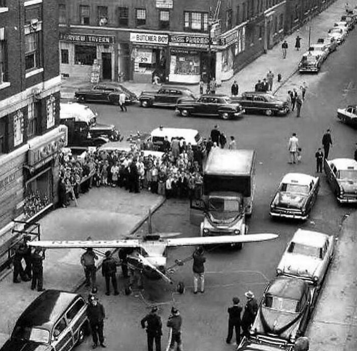 Historic photo of a small plane on a city street, surrounded by onlookers, showcasing an interesting history moment.