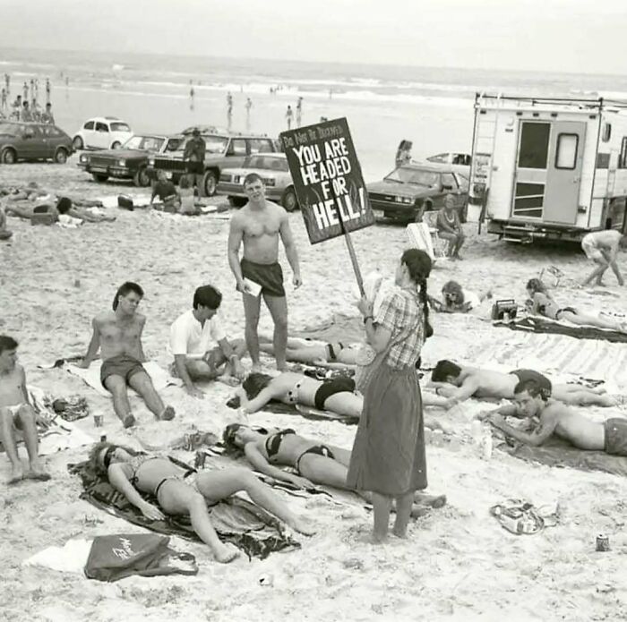 Woman holding sign reading "You are headed for hell" on a crowded beach, capturing a moment from the past.