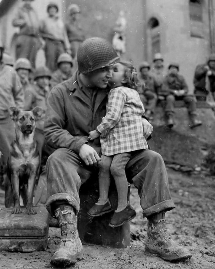 Soldier smiling as a young girl kisses his cheek, with a dog and fellow soldiers in the background, illustrating historical moments.