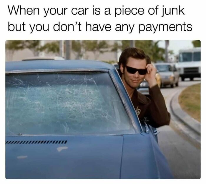 Person in sunglasses smiling, posing by a damaged car, humorously reflecting work meme vibes.