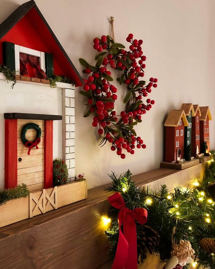 Christmas decorating ideas with a red berry wreath, small festive houses, and glowing garland on a wooden mantel.