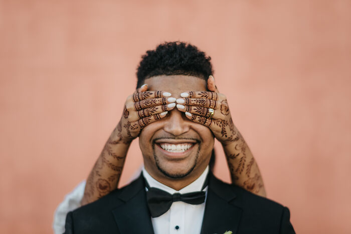 Groom smiling as hands with henna cover his eyes, showcasing creative wedding photography.