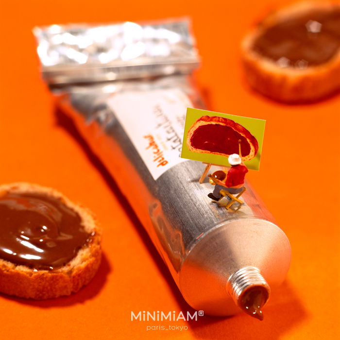 Miniature artist painting food on a tiny art setup beside a tube with spread and bread, showcasing creativity in tiny worlds.