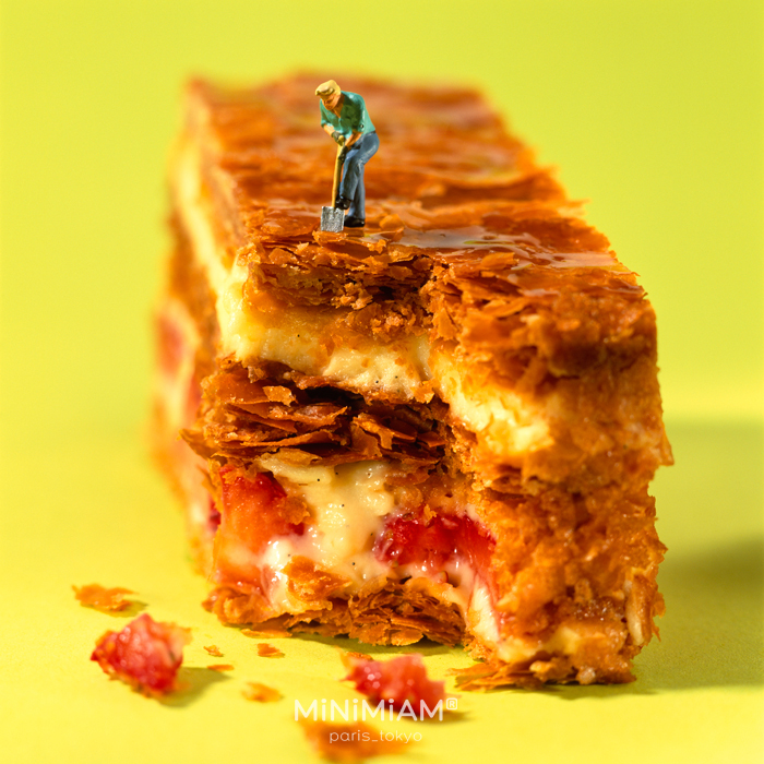 Miniature worker digging into a pastry, highlighting the tiny world of creative food miniatures on a yellow background.