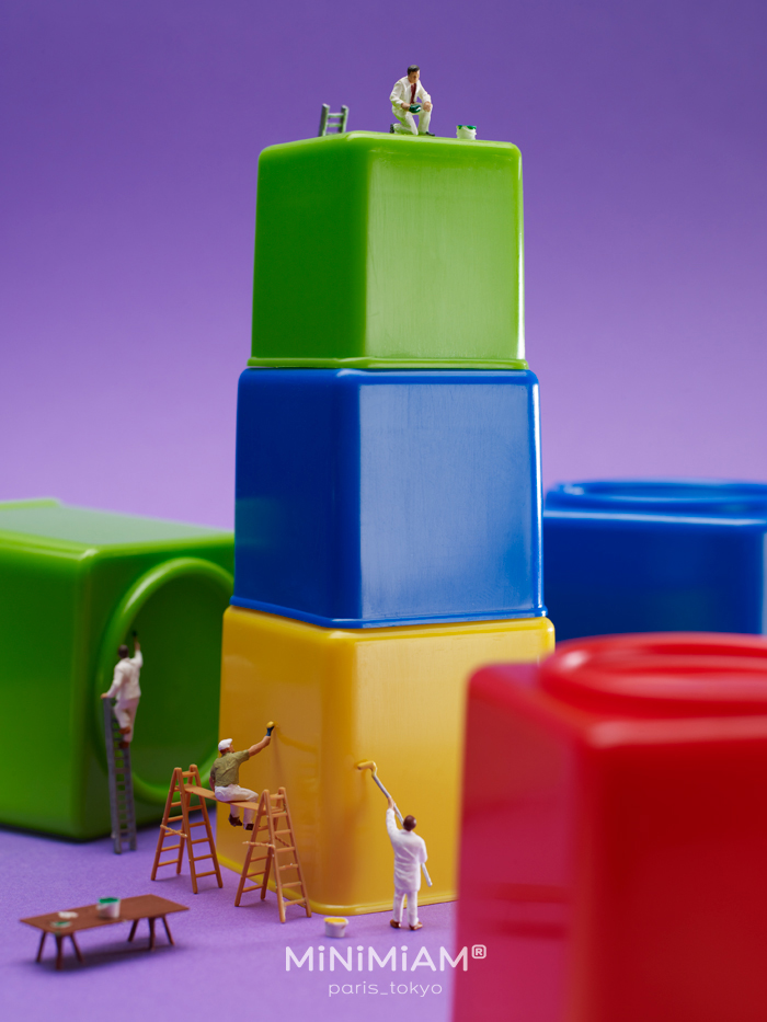 Miniature figures painting colorful blocks, showcasing creative tiny world art on a purple background.