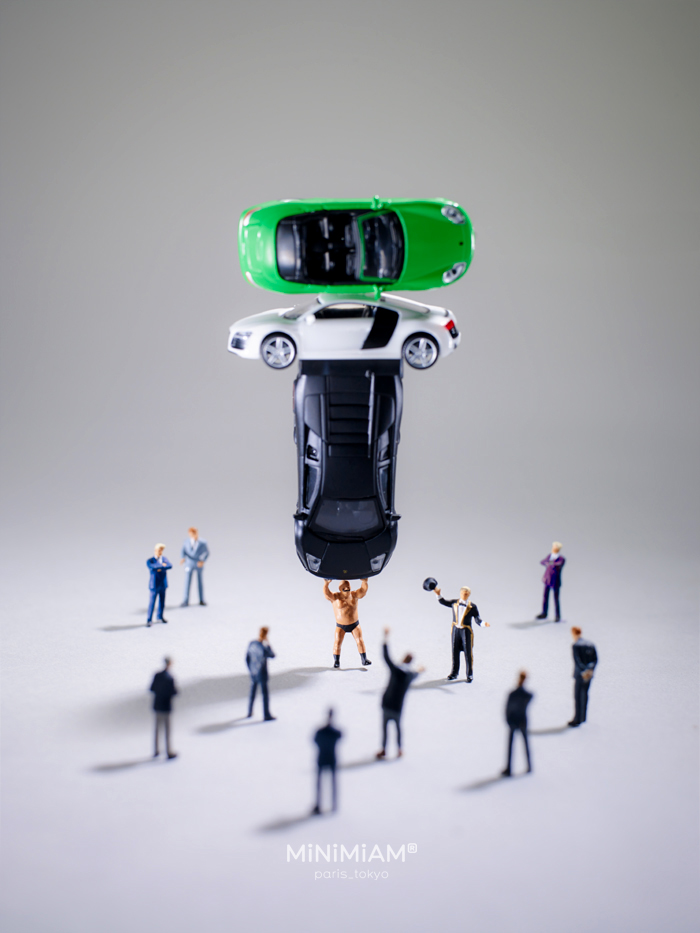 Miniatures of people surrounding a small figure lifting stacked toy cars, showcasing a tiny world of creative scenes.