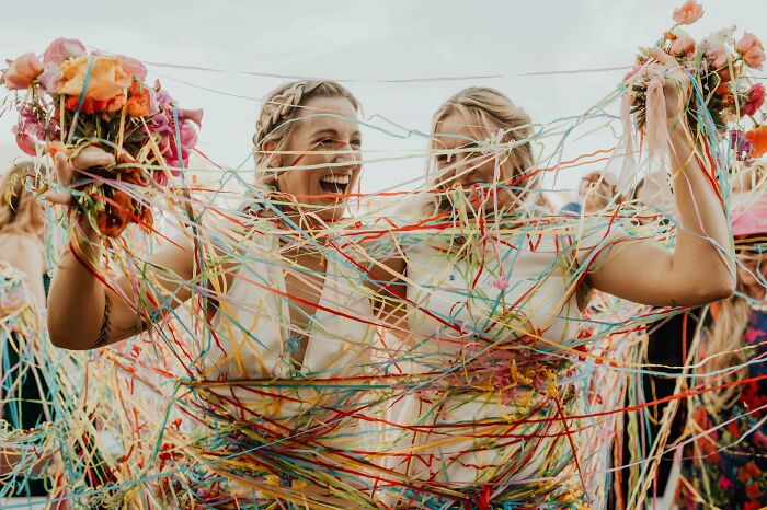 Two brides laughing, covered in colorful streamers, holding flower bouquets at a vibrant wedding celebration.