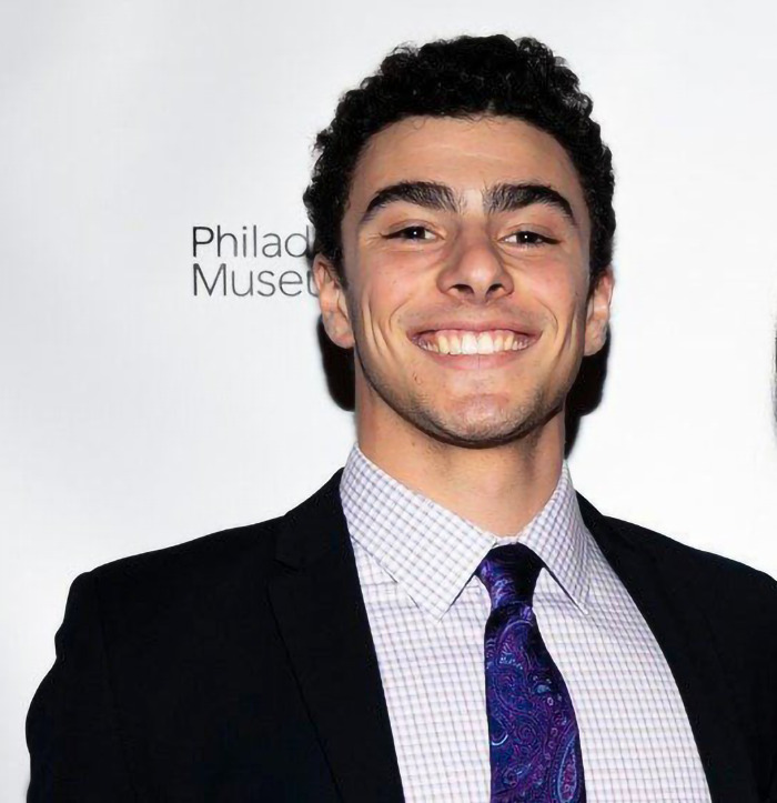 Luigi Mangione in a suit, smiling against a white background. 