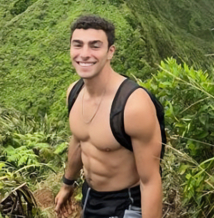 Luigi Mangione smiling on a hiking trail with lush green hills in the background.