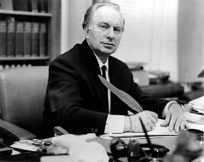 Scientology founder L. Ron Hubbard sitting at a desk with a feather pen in hand, wearing a suit, related to ex-Scientologist brainwashing claims.