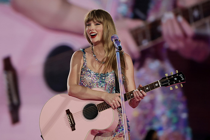 Taylor Swift smiling on stage with a pink guitar, performing live in a sparkling outfit.