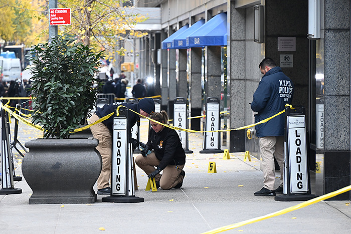 Police investigating crime scene outside a building; yellow tape and evidence markers visible, related to UnitedHealthcare CEO incident.