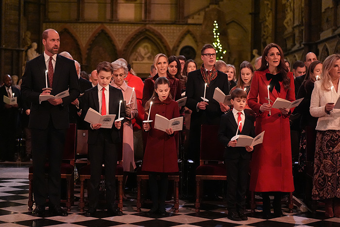 Royal family at Christmas service holding candles, children in red outfits.