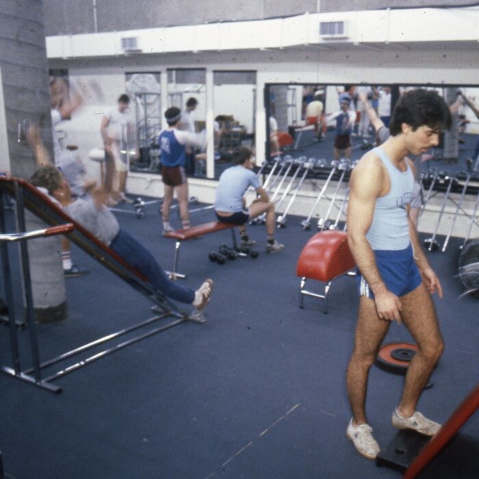 Men exercising in an 80s gym, wearing retro fitness attire, surrounded by workout equipment and mirrors.