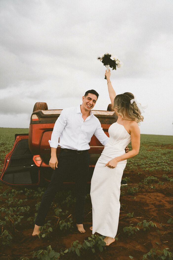 Couple poses joyfully in front of overturned car after accident on way to photo shoot, capturing a unique moment.