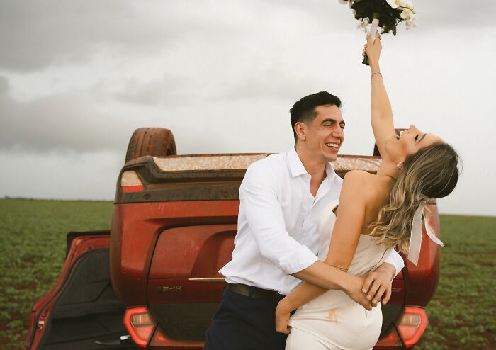 Couple laughing and posing with overturned car after accident, bride lifting bouquet joyfully in field.