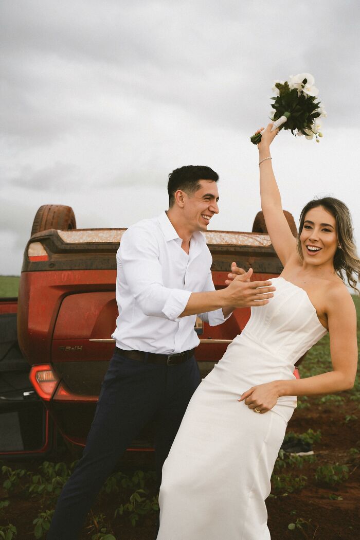 Couple poses joyfully in wedding attire beside overturned car after accident on the way to their photo shoot.