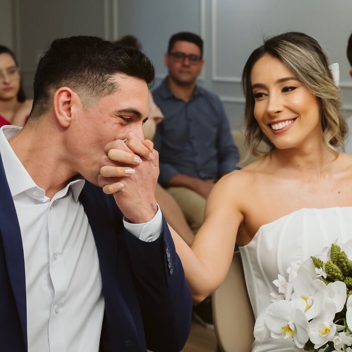 Couple smiling and holding hands, wearing wedding attire, surrounded by people at an event.