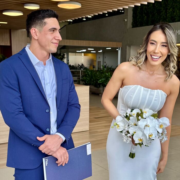 Couple in formal attire smiling indoors, holding flowers, before taking photos at a unique accident scene.