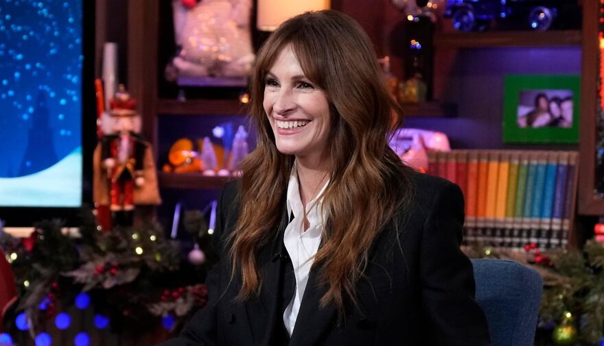 Smiling actor Julia Roberts in a black jacket, sitting indoors with colorful decorations in the background.