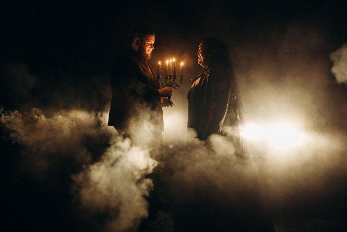 Couple in dark setting holding candelabra amidst smoke, capturing one of 2024's top wedding photo moments.