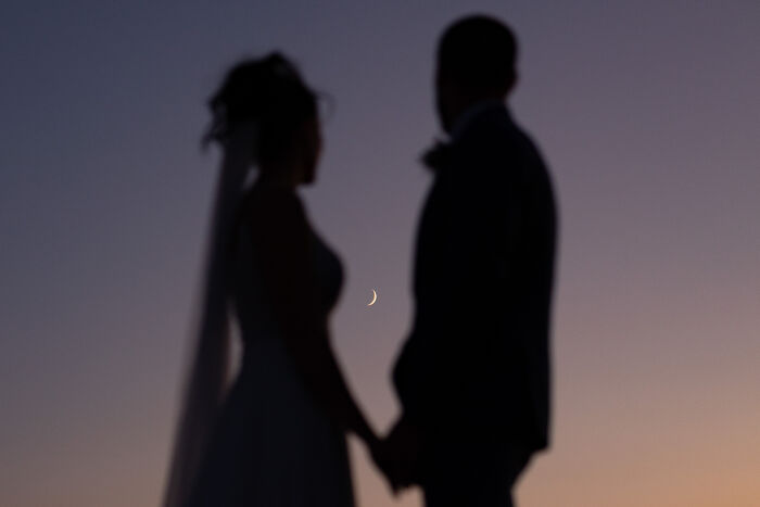 Silhouette of a bride and groom holding hands under a crescent moon at sunset, top wedding photo of 2024.