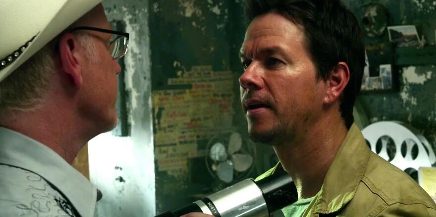 Two actors Mark Wahlberg in a tense scene, with one holding a microphone towards the other, surrounded by a vintage-looking room.