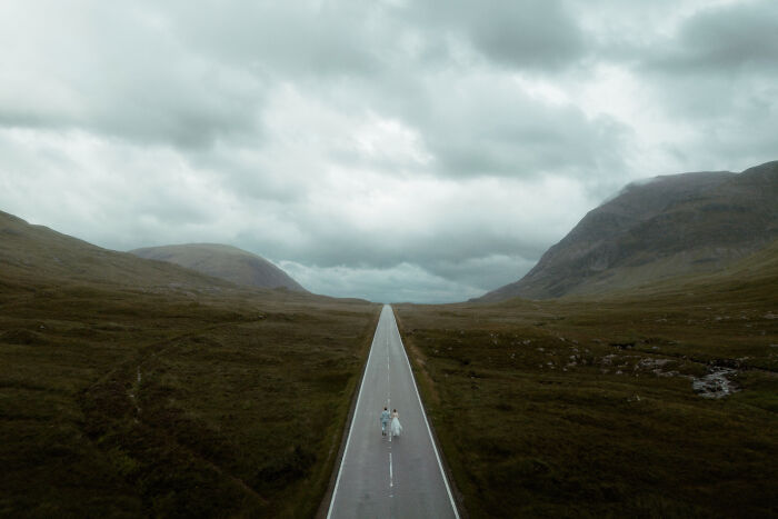 Bride and groom walking on an empty road surrounded by mountains under cloudy skies, from top wedding photos of 2024.