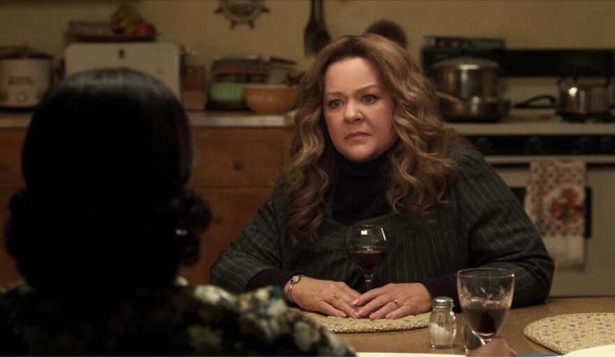 Actor Melissa McCarthy at a dining table, holding a wine glass, in a kitchen setting.