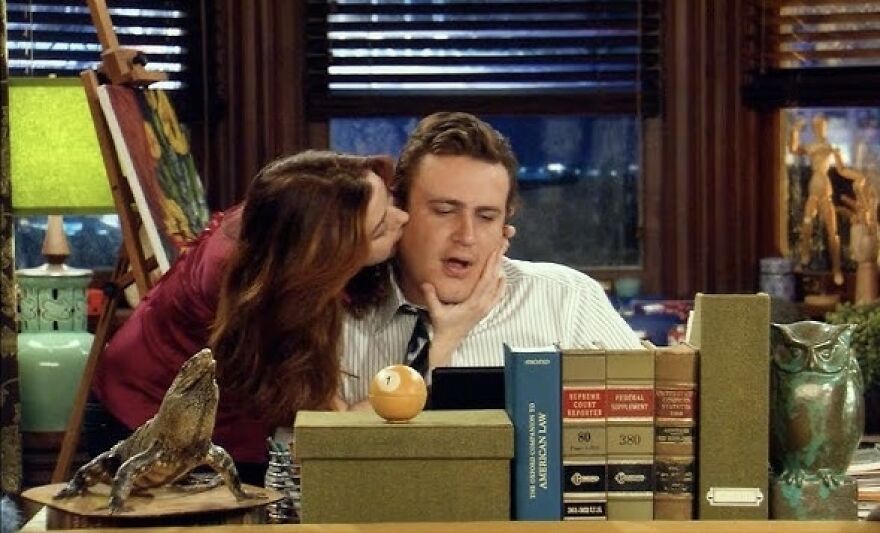 Couple sitting at a desk with one kissing the other on the cheek, surrounded by books and decor in How I Met Your Mother.