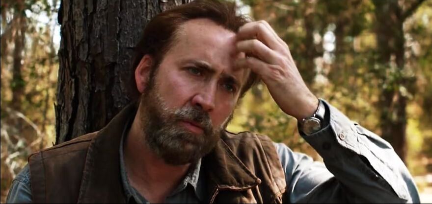 Actor Nicholas Cage with a beard leaning against a tree, wearing a jacket, embodying familiar character traits of actors who play themselves.