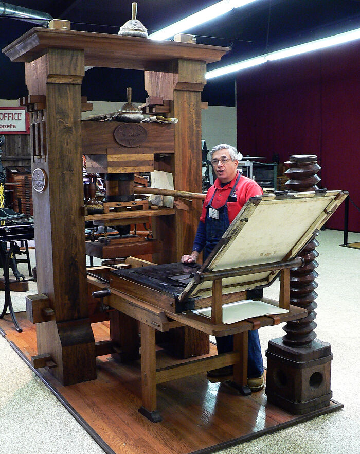 A man stands beside a large wooden printing press, showcasing historical influence on media and technology.