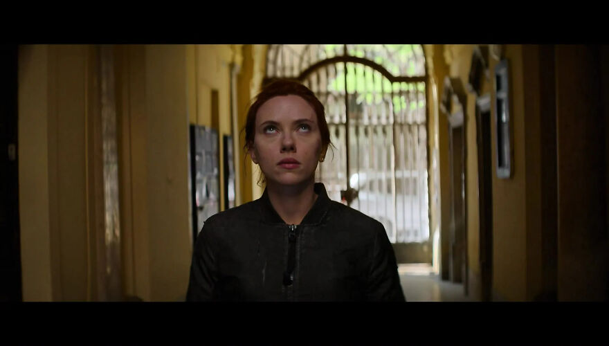 Actress Scarlett Johansson with red hair stands in a dimly lit corridor, looking upwards, wearing a black jacket.