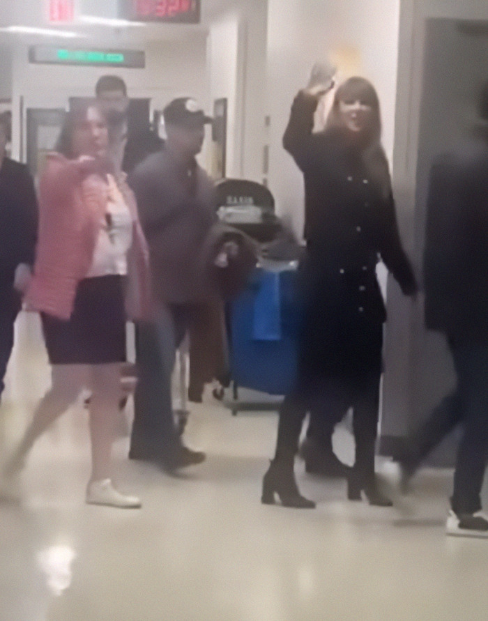 Taylor Swift in a black outfit walking through a hospital corridor, waving to hospital staff.