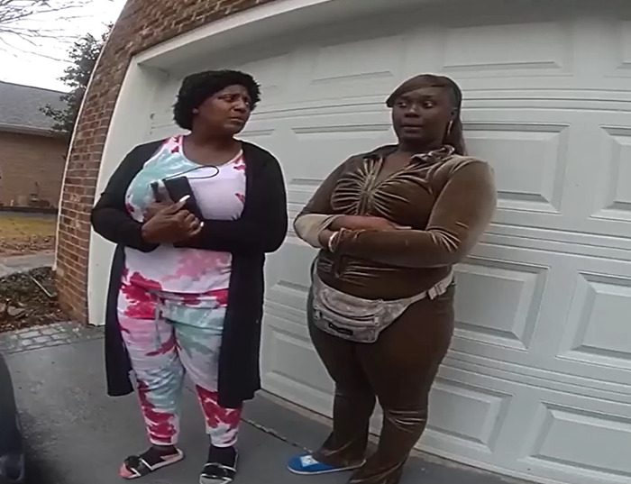 Two women standing outside a garage, discussing a home dispute involving a squatter.