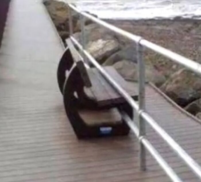 Construction fail: A wooden bench is humorously installed sideways under a boardwalk railing by the beach.