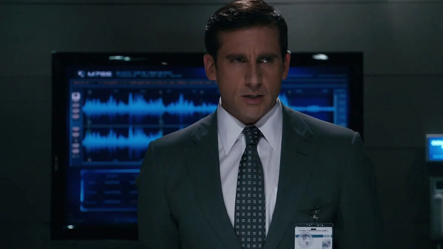 Actor Steve Carell in a suit stands in front of a digital display screen, embodying a role typical of his style.