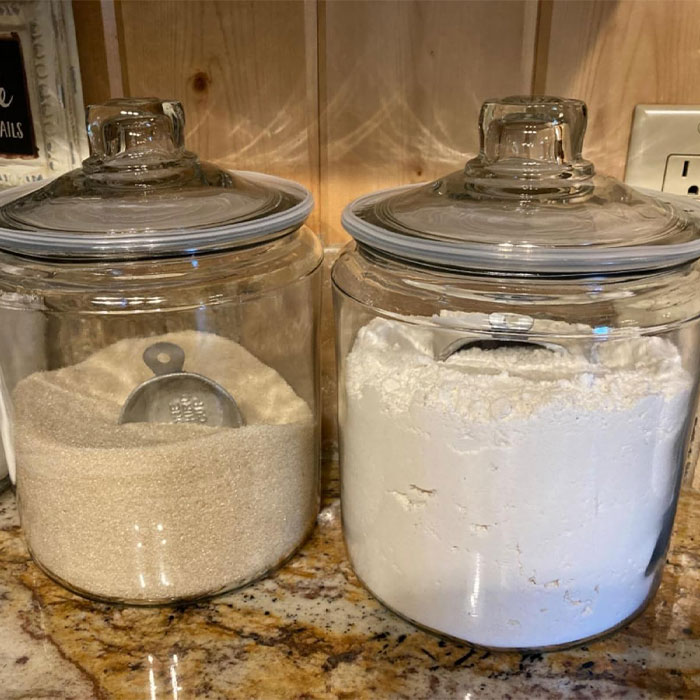 Two glass jars on a countertop, one filled with sugar and the other with flour, both containing measuring cups.