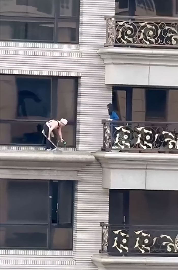 Person cleaning windows on a building ledge while another person watches from a balcony, illustrating humans of capitalism pictures.