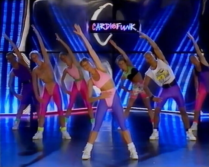 Group performing 80s gym fitness routine in vibrant workout attire, stretching under "Cardiofunk" neon lights.