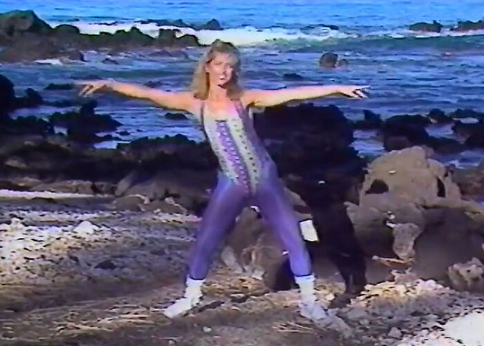 Person in retro 80s gym fitness outfit, exercising on a rocky beach with ocean waves in the background.