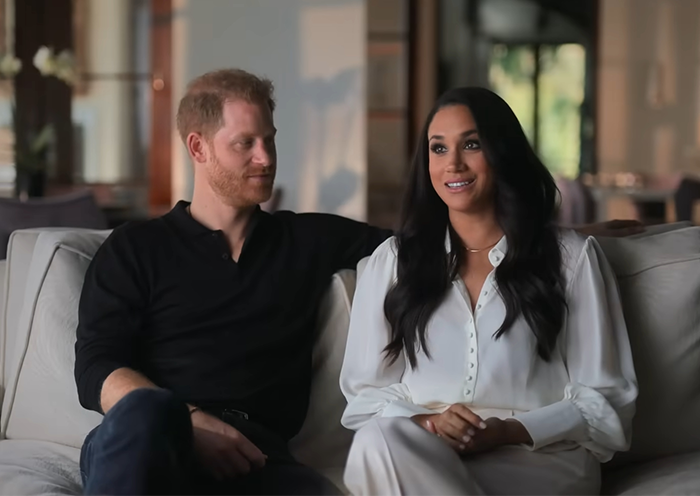 Prince Harry and Meghan Markle sitting on a sofa, both smiling casually in a relaxed setting.