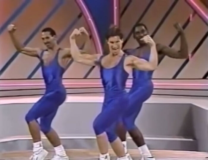 Three men in blue leotards posing energetically in a retro 80s gym fitness setting.