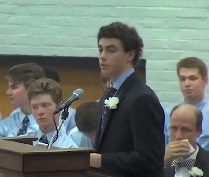 Luigi Mangione speaking at a lectern, wearing a suit with a white flower pinned, surrounded by attentive listeners.