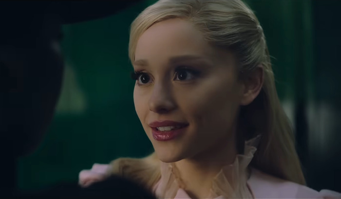 Ariana Grande as Glinda, facing a dark-clad person in a dimly lit setting from movie 'Wicked'.