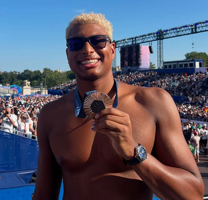 Olympian in Paris holding a rusted bronze medal, smiling at a crowded outdoor event.