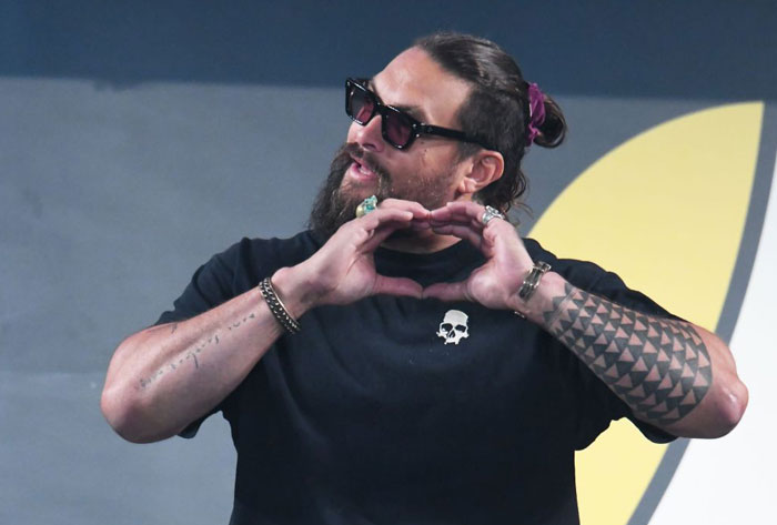 Man with sunglasses forming heart shape, wearing a black shirt with skull logo, representing Aquaman 2 cast.