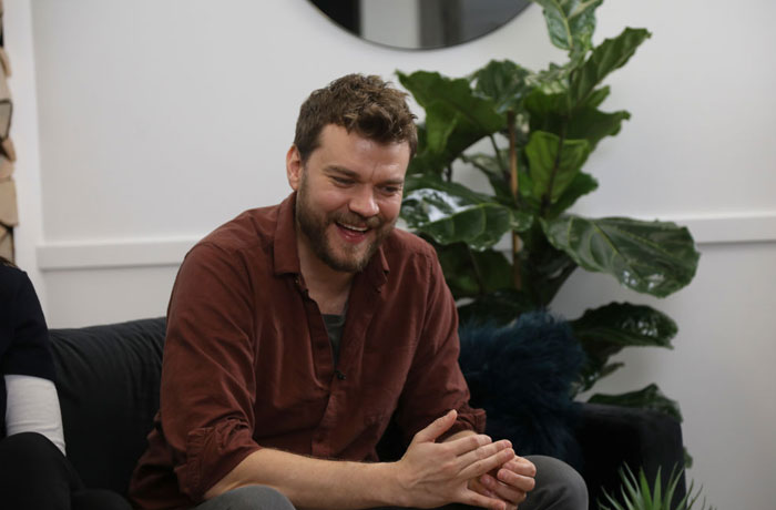 Man in a casual setting, sitting and smiling, with plants in the background, related to Aquaman 2 cast.