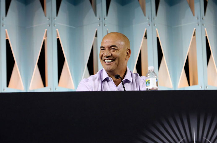 Smiling man at panel discussion related to Aquaman 2 cast, seated with a microphone and water bottle.