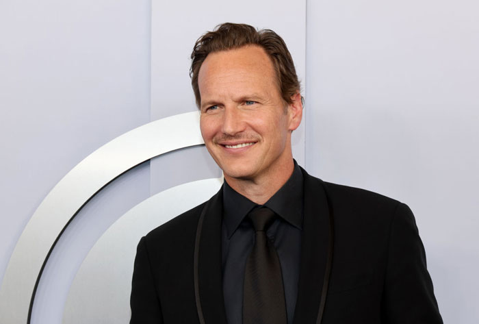 Smiling man in a black suit from Aquaman 2 cast, standing against a light background.